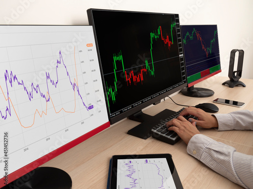 Caucasian young woman sitting at a table doing stock trading on a computer with three black monitors in a white room