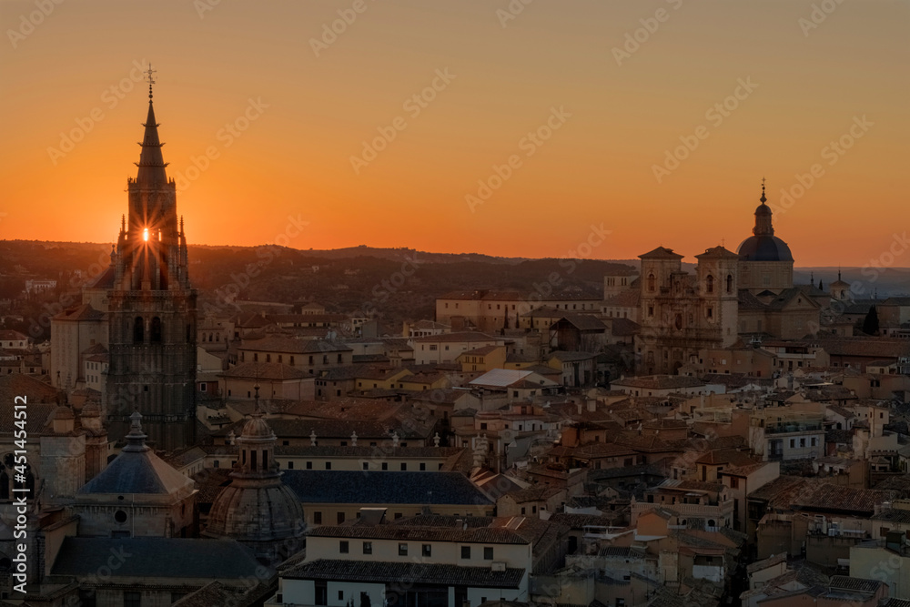 Panoramic view of old part of the city with a sun star between the belfry of the tower of the old cathedral of Toledo in orange sunset, Spain.
