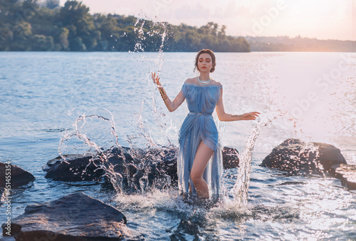 Greek mythical fairytale fantasy woman goddess nymph emerges from lake. Splashes of water. Vintage blue long sexy wet dress. summer nature forest, water surface of river coastal stones. Girl mermaid