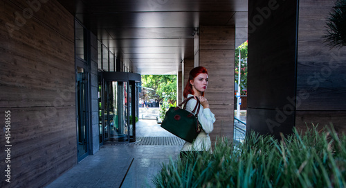 beautiful redhead girl in stylish clothes and a bag near the business building  © Roman