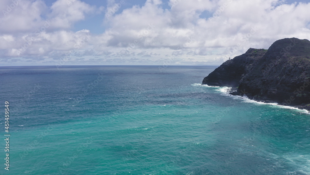 Flying drone over the ocean. View of makapuu lighthouse. Waves of Pacific Ocean wash Rocky shore. Magnificent mountains of Hawaiian island of Oahu against backdrop of blue sky with white clouds.