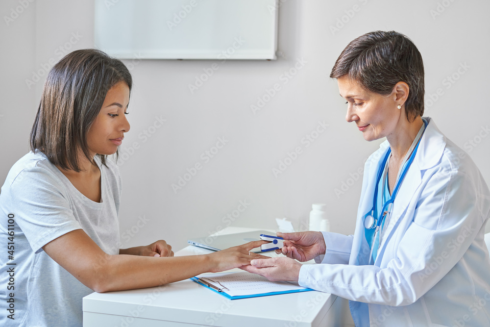 Mature female professional doctor with stethoscope on shoulders measuring oxygen saturation of young afro american woman, patient with pulse oximeter during medical healthcare checkup in clinic office Mature female professional doctor with stethoscope on shoulders measuring oxygen saturation of young afro american woman, patient with pulse oximeter during medical healthcare checkup in clinic office