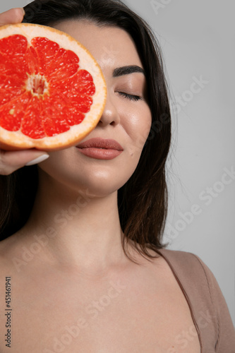 Close-up portrait of a young woman with clean skin holding near face grapefruit on grey background studio portrait.