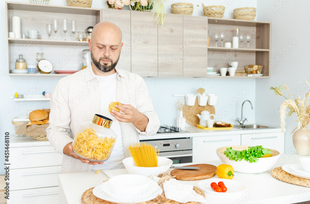 happy young, handsome, bearded man is standing in the modern kitchen with pasta in his hands and vegetables on table, cooking as concept of a man's hobby