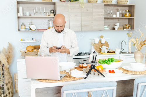 happy young, handsome, bearded man is standing in modern kitchen prepares salad fresh vegetables lettuce, tomatoes, sweet pepper with knife on a cutting board, records video for food blog with laptop