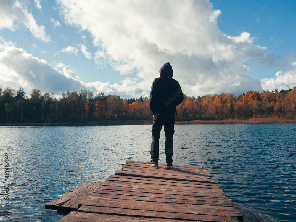 Rear view, a man on a wooden bridge by the lake against the background of an autumn forest.