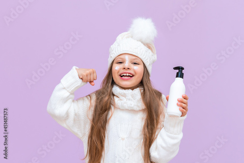 A little girl is holding a cream that protects the skin from winter frosts on an isolated pink background.