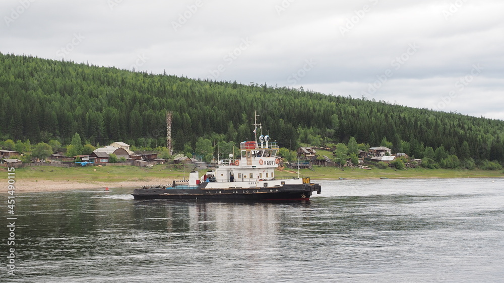 View of Lena river in Ust-Kut city, Siberia. A tugboat ship Nakat owned by Rosmorrechflot, with rural houses in background