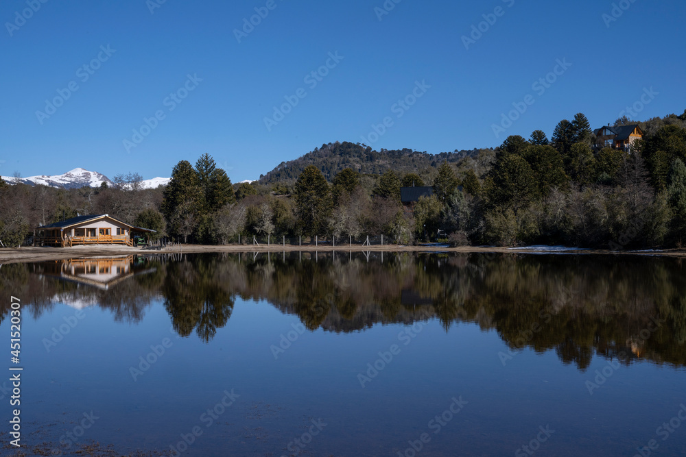 The lake house in the forest. Beautiful view of the summer cabin, calm lake, mountains and forest under a blue sky. The perfect reflection in the water surface. 