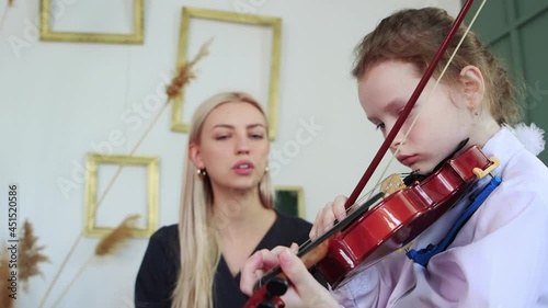Violin lesson - a little girl playing violin with a bow in the class and her teacher sitting next to her and talking