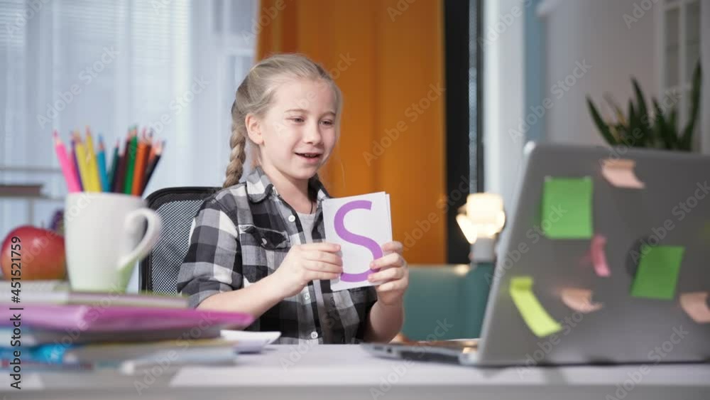 kid girl school pupil does homework using webcam on laptop and shows teacher online cards with letters on computer screen while sitting at table at home kid girl school pupil does homework using webcam on laptop and shows teacher online cards with letters on computer screen while sitting at table at home