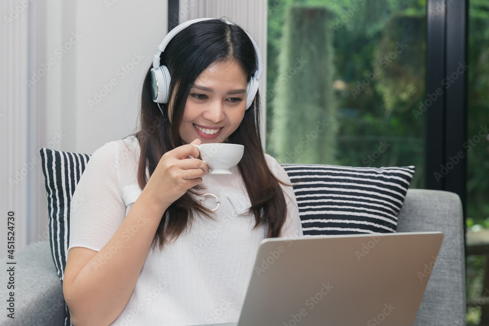 Pretty smiling young woman wear headphones sitting on sofa holding a cup of coffee and usinging laptop. Pretty smiling young woman wear headphones sitting on sofa holding a cup of coffee and usinging laptop.