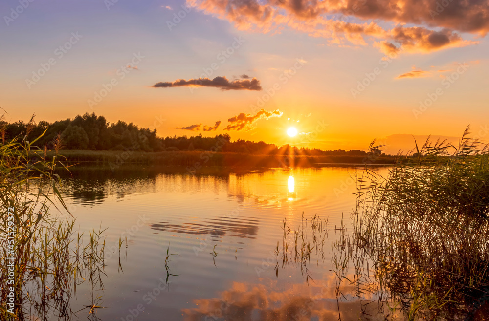 Scenic view at beautiful summer sunset on lake with reflection on water with reeds, grass, golden sun rays, calm water ,deep blue cloudy sky and glow on a background, spring evening landscape
