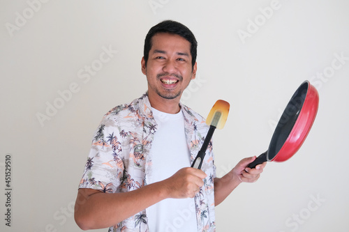 Cheerful Asian man holding frying pan and spatula when cooking in the kitchen photo