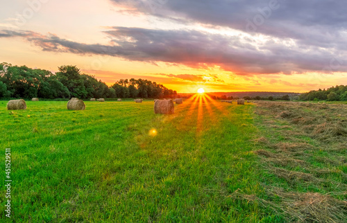 Scenic view at sunset or sunrise in green shiny field with hay stacks, bright cloudy sky, golden sun rays, summer valley landscape