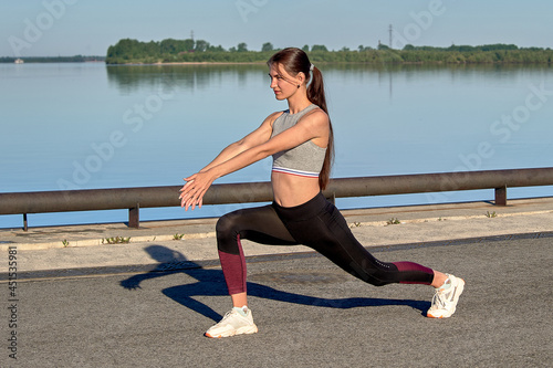 A young woman does a warm-up on the pier in the early morning, against the background of the river