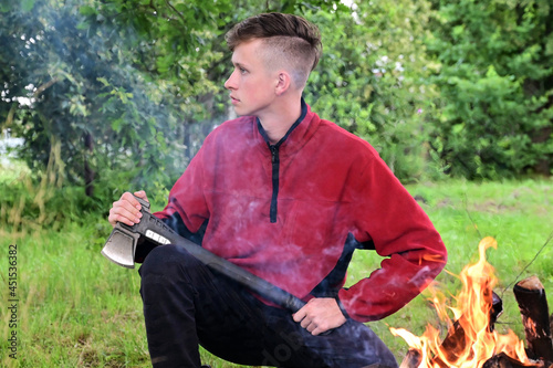 young serious man with a mohawk hairstyle is sitting with an axe by a fire in the forest. Take a break. Path to nature. Concept of adventure photo