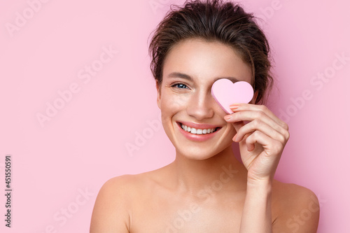 Smiling woman with cosmetic sponge covering one eye. Photo of woman with perfect makeup on pink background. Beauty concept
