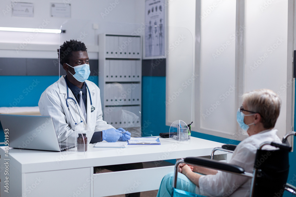 Obraz premium Caucasian patient with disability receiving consultation from african american doctor in medical cabinet. Invalid elder woman sitting in wheelchair for appointment during pandemic