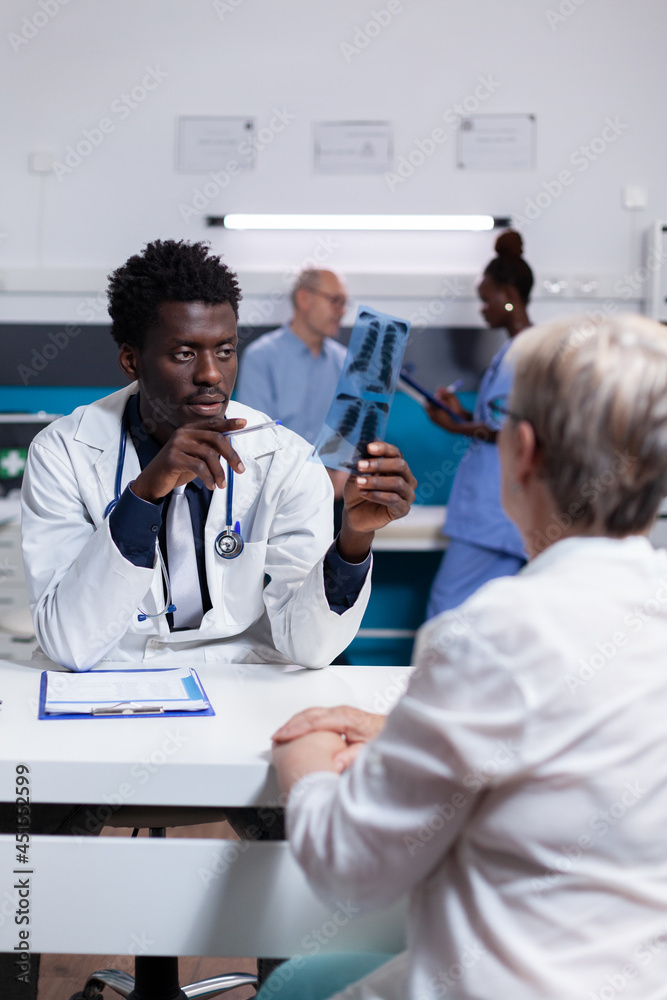 Medic of african american ethnicity holding x ray scan explaining disease to elder woman in cabinet. Black man and old patient doing checkup while nurse and senior man talking in background Medic of african american ethnicity holding x ray scan explaining disease to elder woman in cabinet. Black man and old patient doing checkup while nurse and senior man talking in background