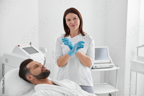 Positive cosmetologist holds syringes with filler for skin lifting standing by mature man in salon