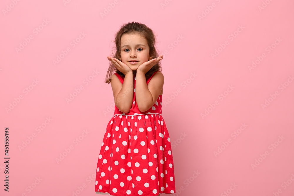 Adorable girl in a pink dress with polka dots poses at the camera, standing against a pink background and holding her chin with her hands. Portrait of a beautiful girl with positive joyful emotions Adorable girl in a pink dress with polka dots poses at the camera, standing against a pink background and holding her chin with her hands. Portrait of a beautiful girl with positive joyful emotions