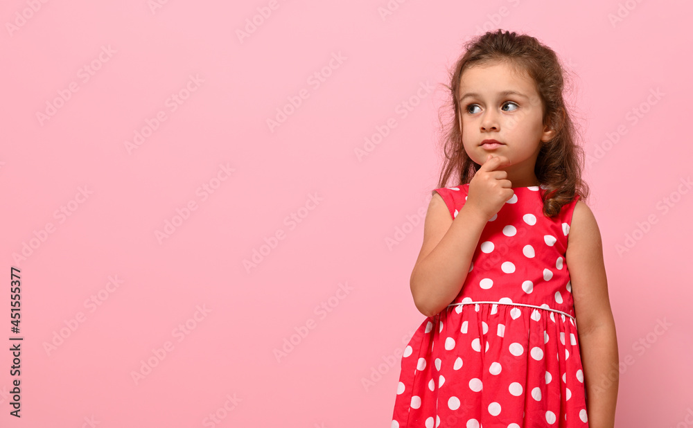 A cute adorable girl of preschool age, 3-4 years old, looks thoughtfully to the side , holding her finger at her chin, posing on a pink background with copy space