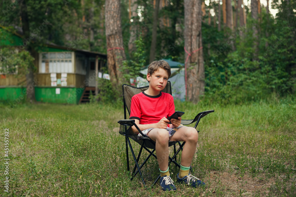 Fototapeta premium boy sitting in a folding chair and holding a smartphone against the background of summer nature
