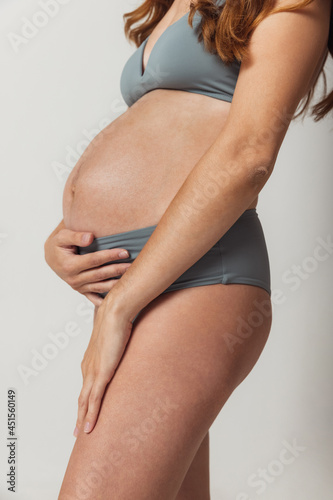 Close-up body, belly of pregnant young woman in lingerie isolated over grey studio background. Natural beauty, happy motherhood, femininity concept.