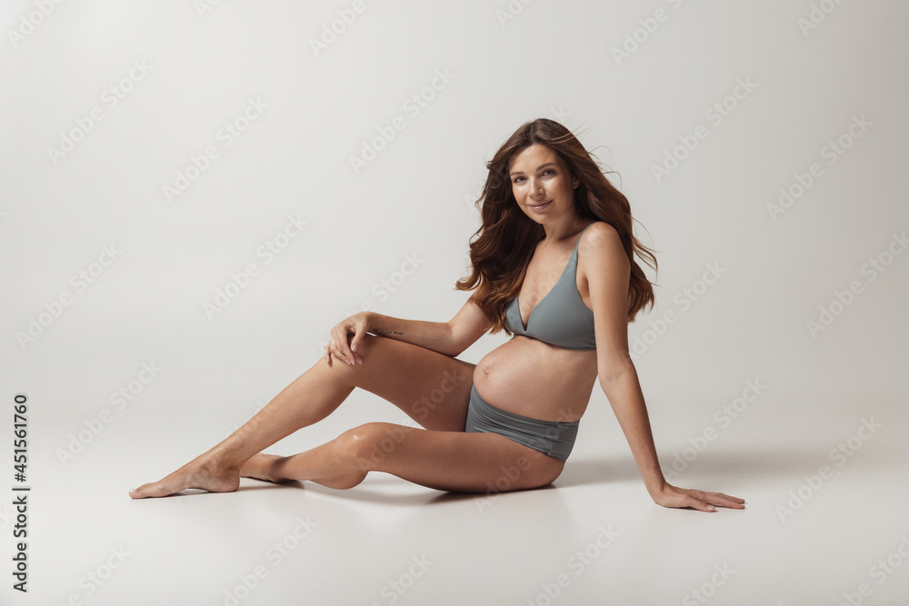 Happy time, awaiting. Portrait of smiling beautiful pregnant woman in lingerie isolated over grey studio background. Natural beauty, happy motherhood, femininity concept. Happy time, awaiting. Portrait of smiling beautiful pregnant woman in lingerie isolated over grey studio background. Natural beauty, happy motherhood, femininity concept.