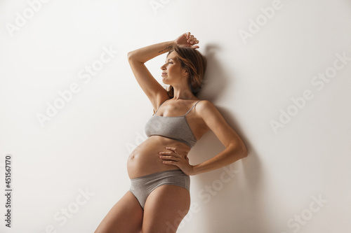 Happy time, awaiting. Portrait of smiling beautiful pregnant woman in lingerie isolated over grey studio background. Natural beauty, happy motherhood, femininity concept.