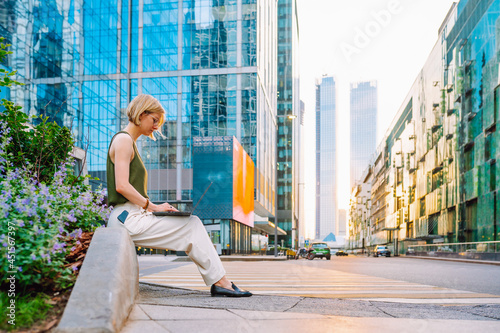 Blonde business woman wearing glasses sit with laptop among the skyscrapers and work.