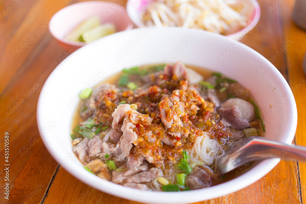 Meatballs and meat with wide rice noodles in soup was added pickled chili on top in a white bowl on woody background. It is a Asian traditional food in Thailand. Meatballs and meat with wide rice noodles in soup was added pickled chili on top in a white bowl on woody background. It is a Asian traditional food in Thailand.