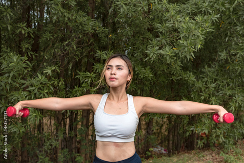 A fit and slender asian lady does standing lateral raises at a nature park. Shoulder workout training. photo