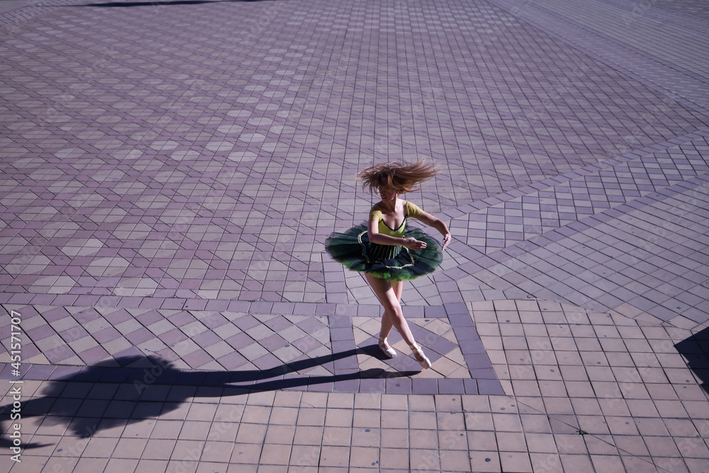 Adult female Hispanic classical ballet dancer in green and black tutu with coins, dancing in the middle of a plaza casting her shadow on the ground. Adult female Hispanic classical ballet dancer in green and black tutu with coins, dancing in the middle of a plaza casting her shadow on the ground.