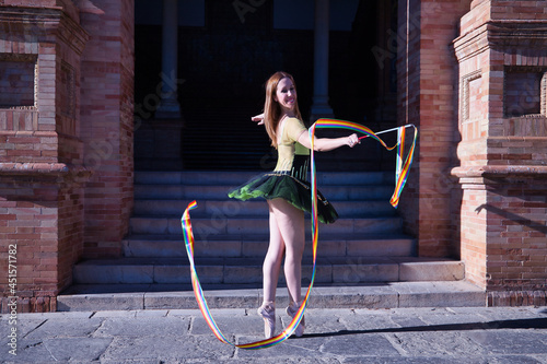 Hispanic adult female classical ballet dancer in green and black tutu with coins, dancing with a rhythmic gymnastics ribbon in the colors of the gay pride flag. © Manuel