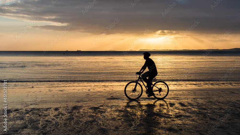 Fototapeta premium Silhouette of a man bike riding on the sand beach at sunrise