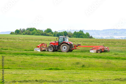 A red tractor mows the grass with two mowers. Harvesting hay for cows for the winter.
