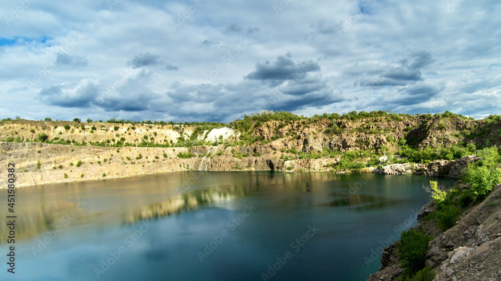 Panoramic view of an old flooded granite quarry against cloudy sky in early summer. Landscape with rock stones, green trees and clean pond
