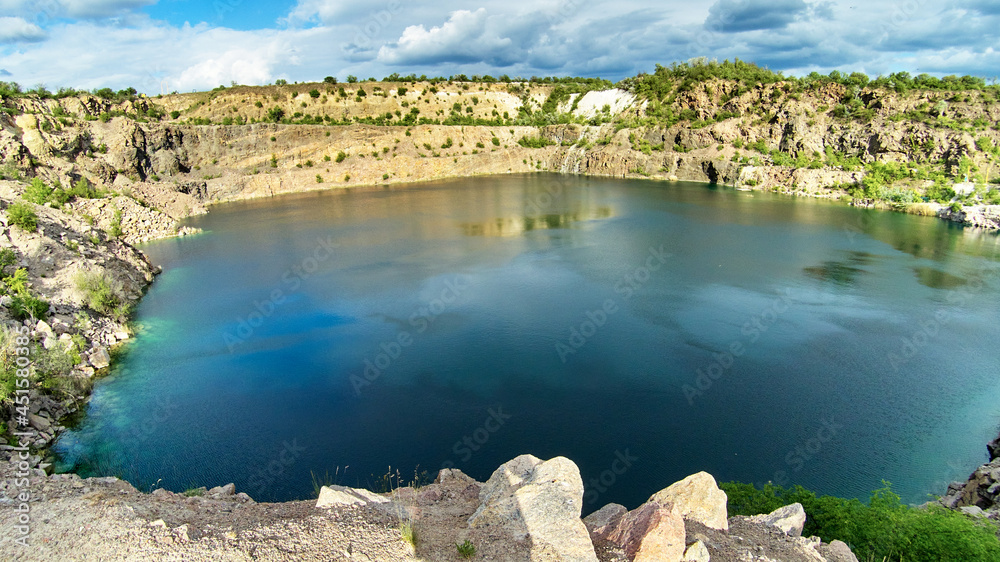 Panoramic view of an old flooded granite quarry against cloudy sky in early summer. Landscape with rock stones, green trees and clean pond