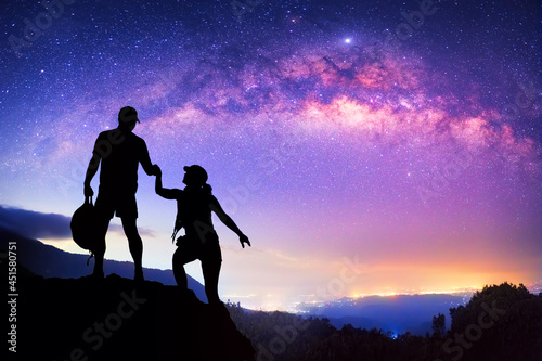 Silhouette young couple climbing mountain against background of colorful starry sky.