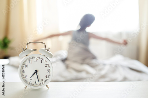 little boy sitting and stretching in bed at home in the morning on a window background with alarm clock