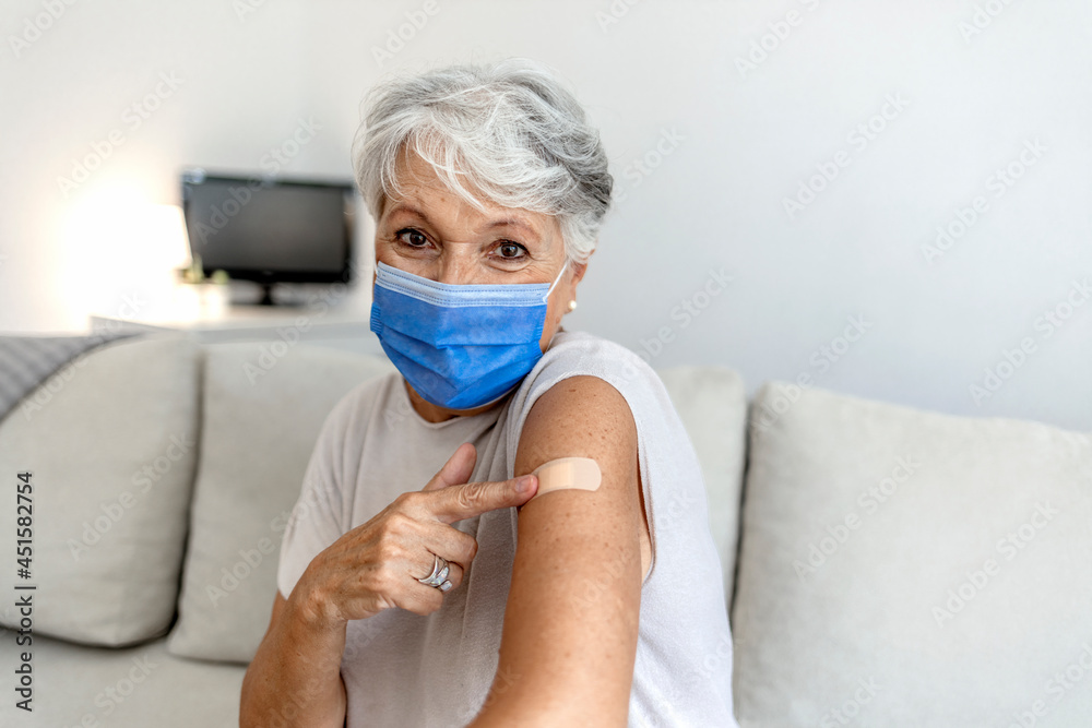 Mature woman against white background after receiving COVID-19 vaccination, wearing protective face mask. Old Caucasian lady holding up shirt sleeve to show the sticking plaster after a flu jab in arm - obrazy, fototapety, plakaty 