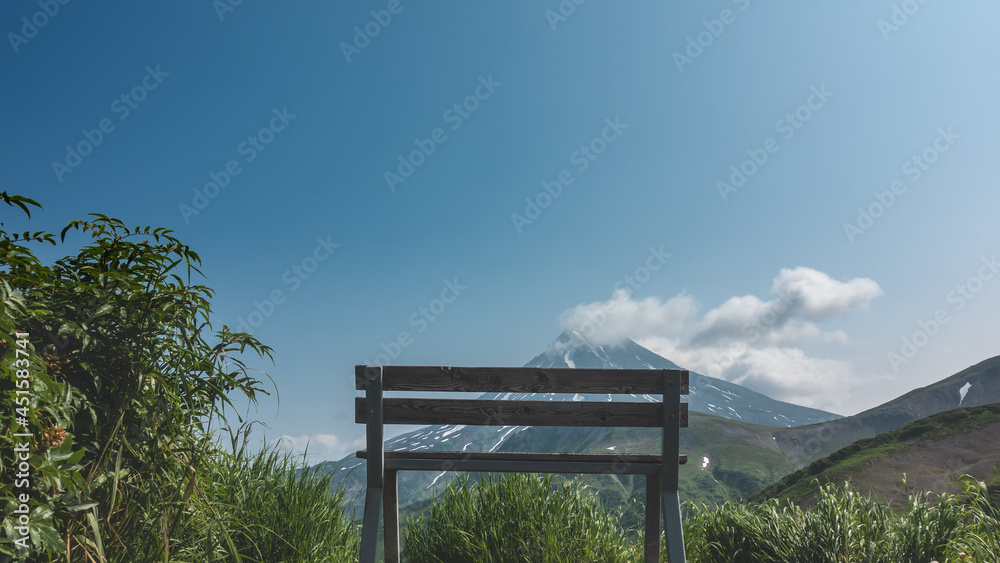 There is an unpainted wooden bench among the green grass. Ahead, against the blue sky, a conical volcano. There is snow on the slopes. Clouds over the top. Kamchatka. Stratovolcano of Vilyuchinsky There is an unpainted wooden bench among the green grass. Ahead, against the blue sky, a conical volcano. There is snow on the slopes. Clouds over the top. Kamchatka. Stratovolcano of Vilyuchinsky