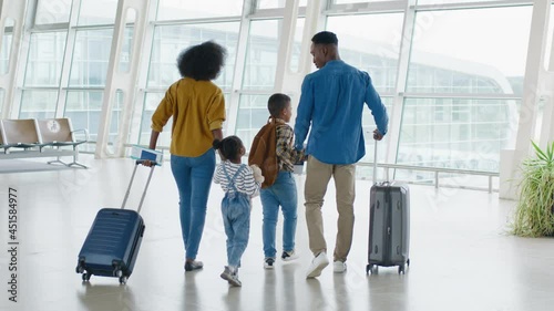 Back view on the multiracial family - mother, father, daughter and son - walking through the airport hall and carrying their suitcases on the wheels before the departure to the vacation