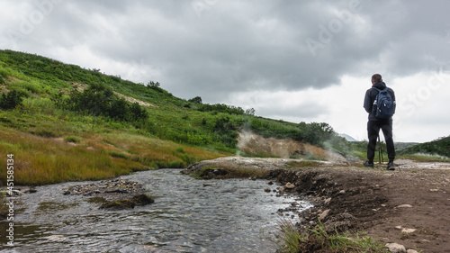 A tourist with a backpack walks along the valley of hot springs along the river. View from the back. Steam jets rise above the ground . There is green vegetation on the hillside. Cloudy. Kamchatka