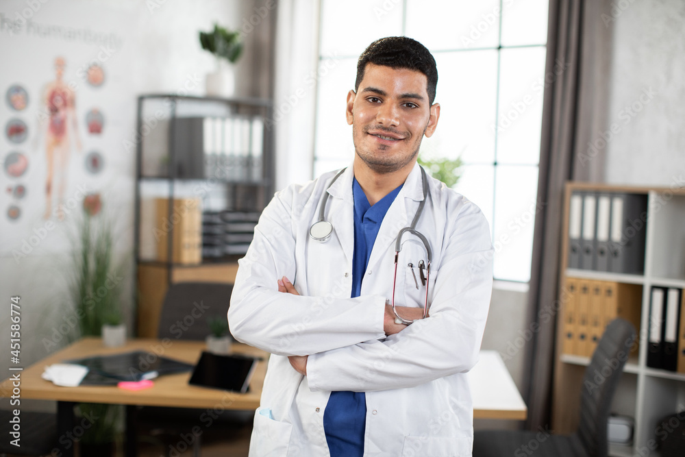 Smiling young 30-aged male Arabic Asian doctor posing with arms crossed in the office. Male physician in blue suit and white coat, with a stethoscope around neck, posing to camera indoors Smiling young 30-aged male Arabic Asian doctor posing with arms crossed in the office. Male physician in blue suit and white coat, with a stethoscope around neck, posing to camera indoors