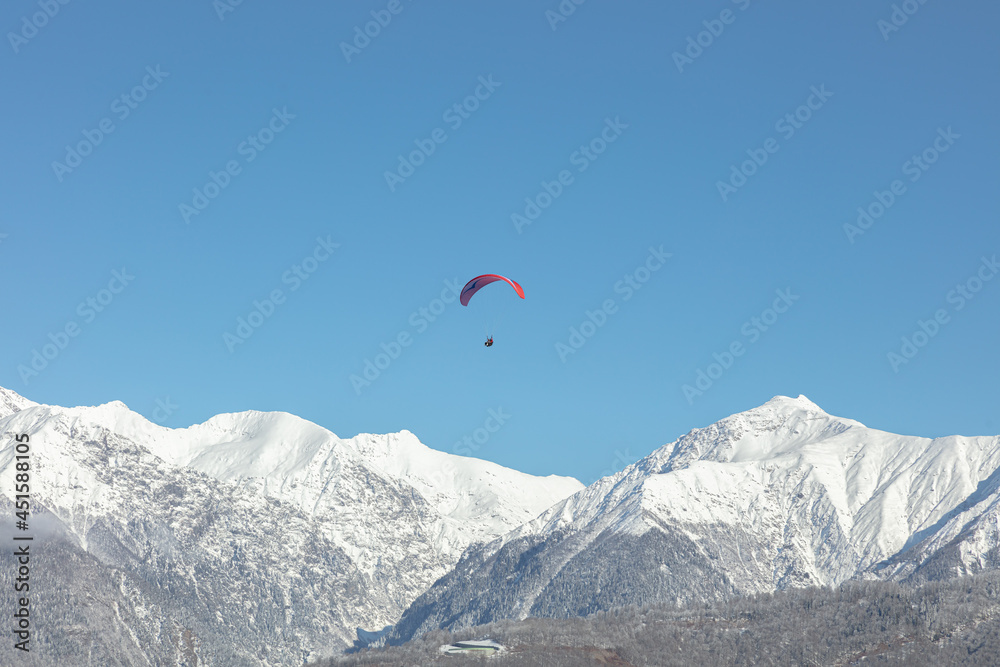 the red wing of the paraglider against the background of the blue sky and the snow-white peaks of the high mountains. the parachutist during the flight against the background of the Alpine mountains the red wing of the paraglider against the background of the blue sky and the snow-white peaks of the high mountains. the parachutist during the flight against the background of the Alpine mountains