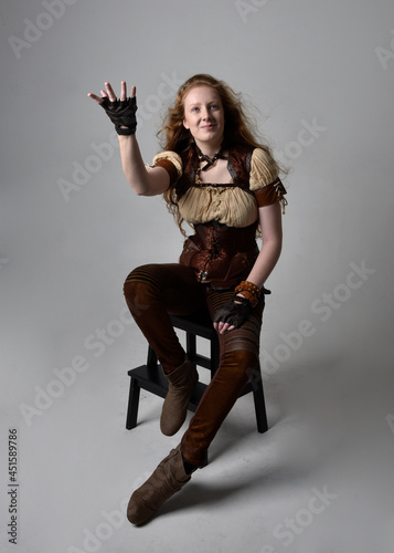 Full length portrait of beautiful young woman with long red hair, wearing steampunk inspired costume. sitting pose isolated on studio background.