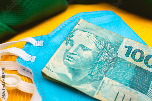 Real - BRL. A pandemia do novo coronavírus e economia brasileira. Uma cédula de 100 reais sobre uma máscara n95 de cor azul. Na composição da foto uma representação da bandeira do Brasil. photo